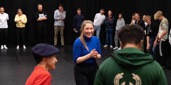 Kvinne i blå skjorte leder aktivitet for ulike grupper mennesker i teaterrommet, med tilskuere som ser på fra bakgrunnen.