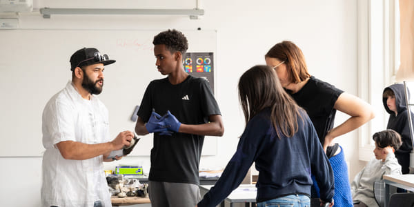 En gruppe ungdommer står rundt et bord og diskuterer en oppgave i et lyst klasserom.