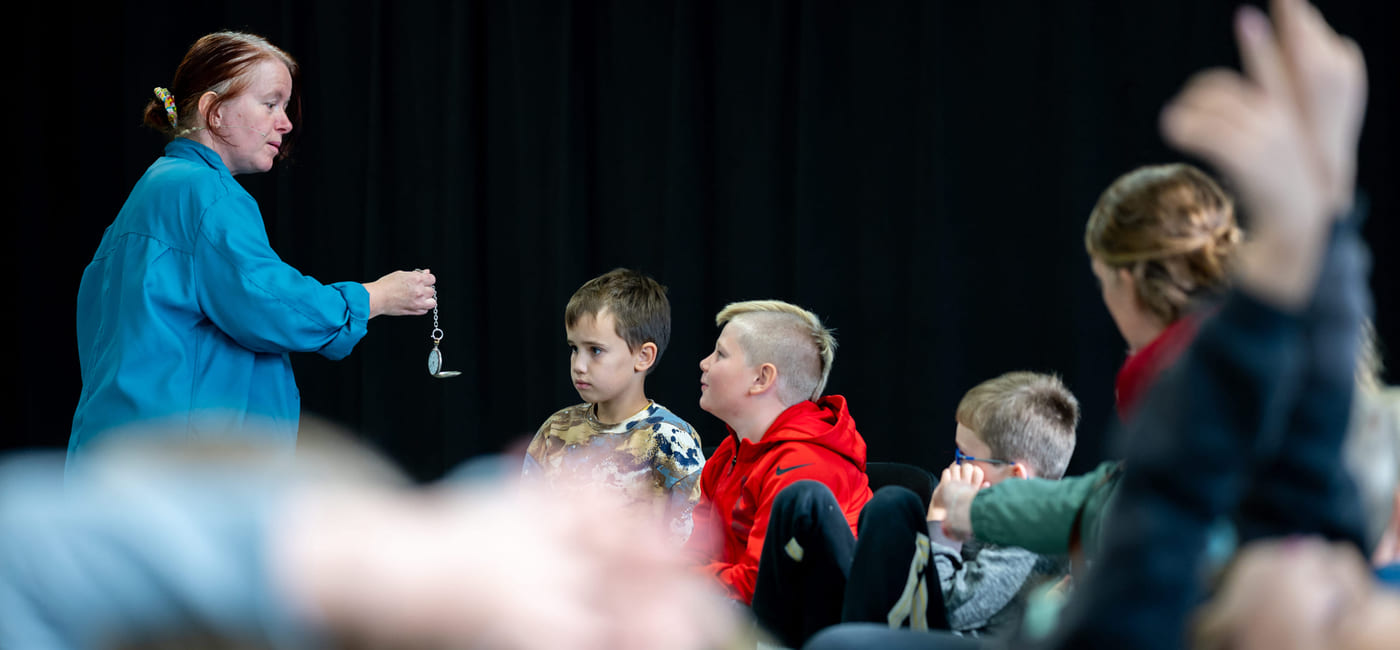 Kvinne i blått demonstrerer lommeur for oppmerksomme barn som sitter i publikum. Ett barn rekker opp hånden i bakgrunnen.