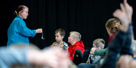 Kvinne i blått demonstrerer lommeur for oppmerksomme barn som sitter i publikum. Ett barn rekker opp hånden i bakgrunnen.