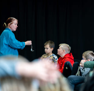 Kvinne i blått demonstrerer lommeur for oppmerksomme barn som sitter i publikum. Ett barn rekker opp hånden i bakgrunnen.