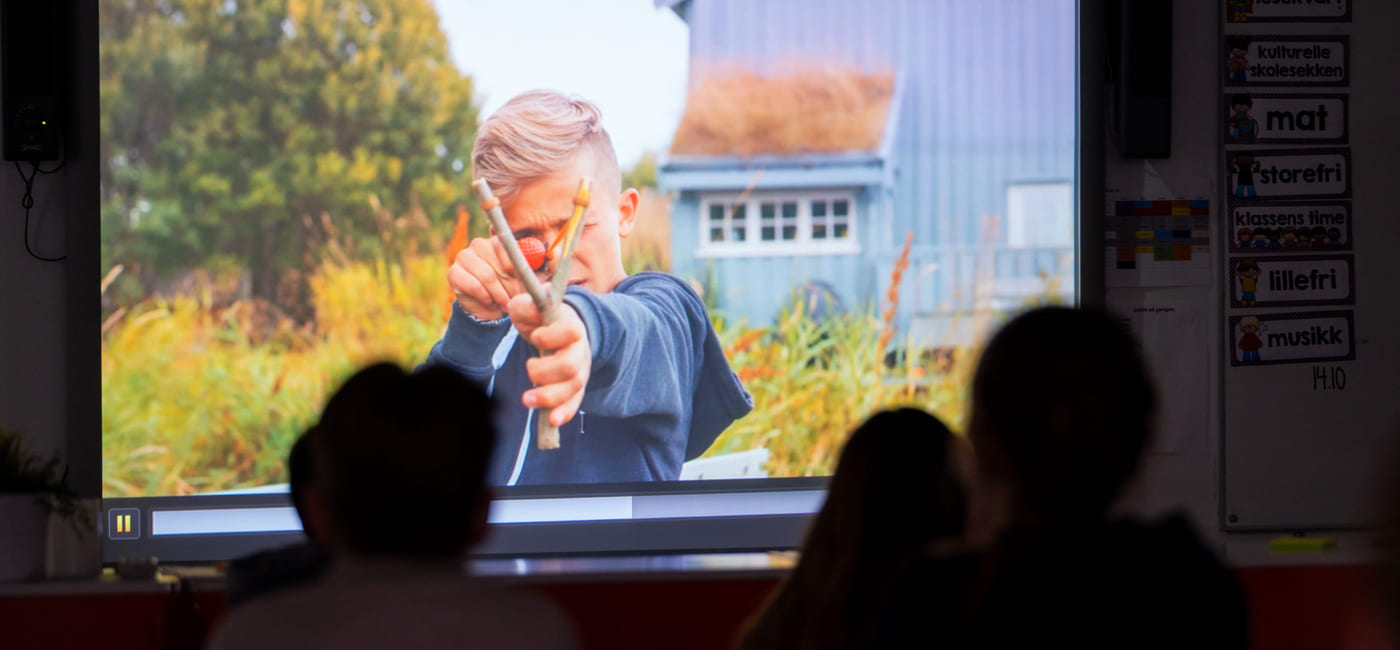 To elever ser på en filmprojeksjon i et klasserom, der en eldre person vises på skjermen foran dem.