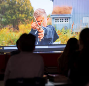 To elever ser på en filmprojeksjon i et klasserom, der en eldre person vises på skjermen foran dem.