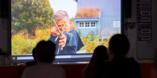 To elever ser på en filmprojeksjon i et klasserom, der en eldre person vises på skjermen foran dem.