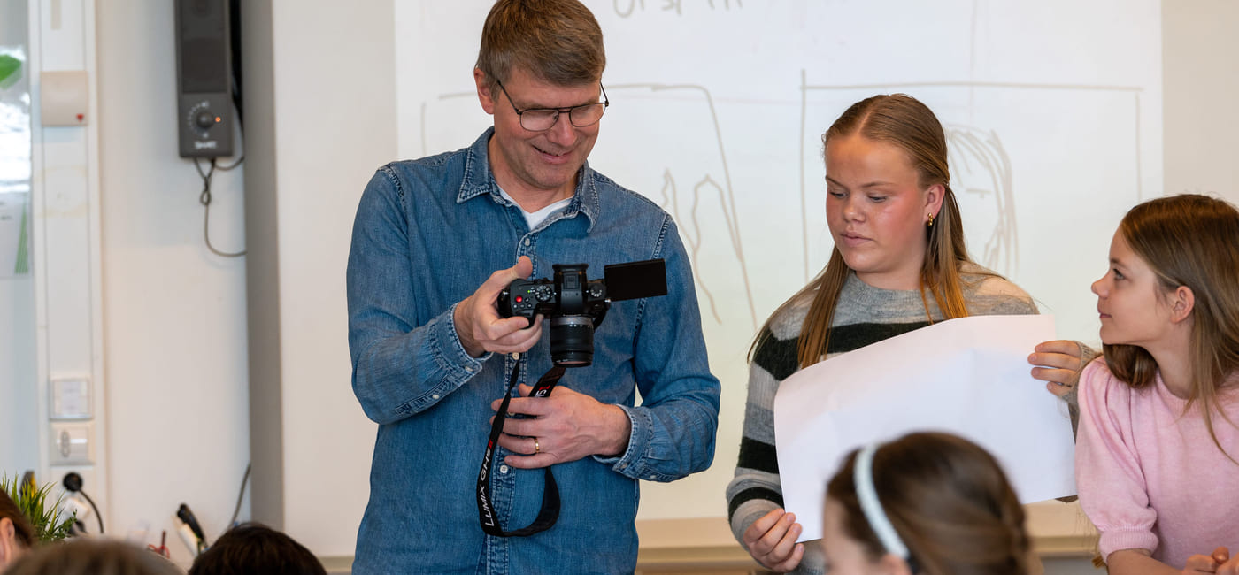 En mann viser fram en gjenstand til en gruppe barn i et klasserom, mens en kvinne står ved siden av og smiler.