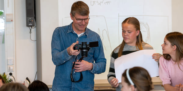 En mann viser fram en gjenstand til en gruppe barn i et klasserom, mens en kvinne står ved siden av og smiler.