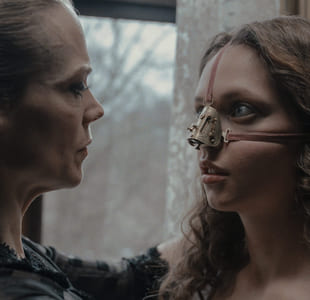 Stillbilde fra filmen "Den stygge stesøsteren". Nærbilde av en kvinne som knytter fast fløyelsbånd rundt ansiktet til ung jente. Båndene holder på plass en buet metallbit med skruer i over nesen til jenta. De ser på hverandre.