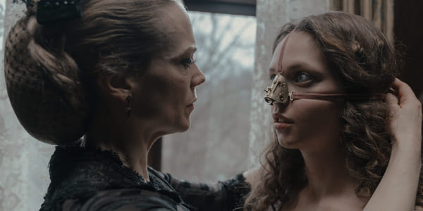 Stillbilde fra filmen "Den stygge stesøsteren". Nærbilde av en kvinne som knytter fast fløyelsbånd rundt ansiktet til ung jente. Båndene holder på plass en buet metallbit med skruer i over nesen til jenta. De ser på hverandre.