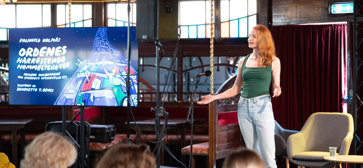 Kvinne med rødt hår presenterer på scenen, publikum ser på. Skjermen viser hendelsestittel på tysk. Innendørs spillested.