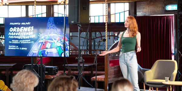 Kvinne med rødt hår presenterer på scenen, publikum ser på. Skjermen viser hendelsestittel på tysk. Innendørs spillested.