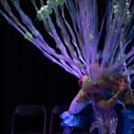Utøvere i glitrende kostymer med livlige lilla og blå tentakellignende forlengelser, som skaper en utenomjordisk undervannseffekt på mørk scene.