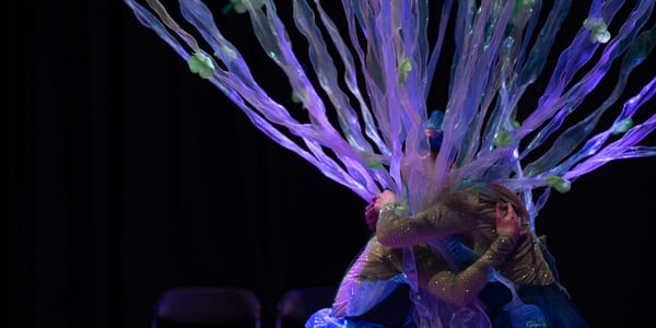 Utøvere i glitrende kostymer med livlige lilla og blå tentakellignende forlengelser, som skaper en utenomjordisk undervannseffekt på mørk scene.