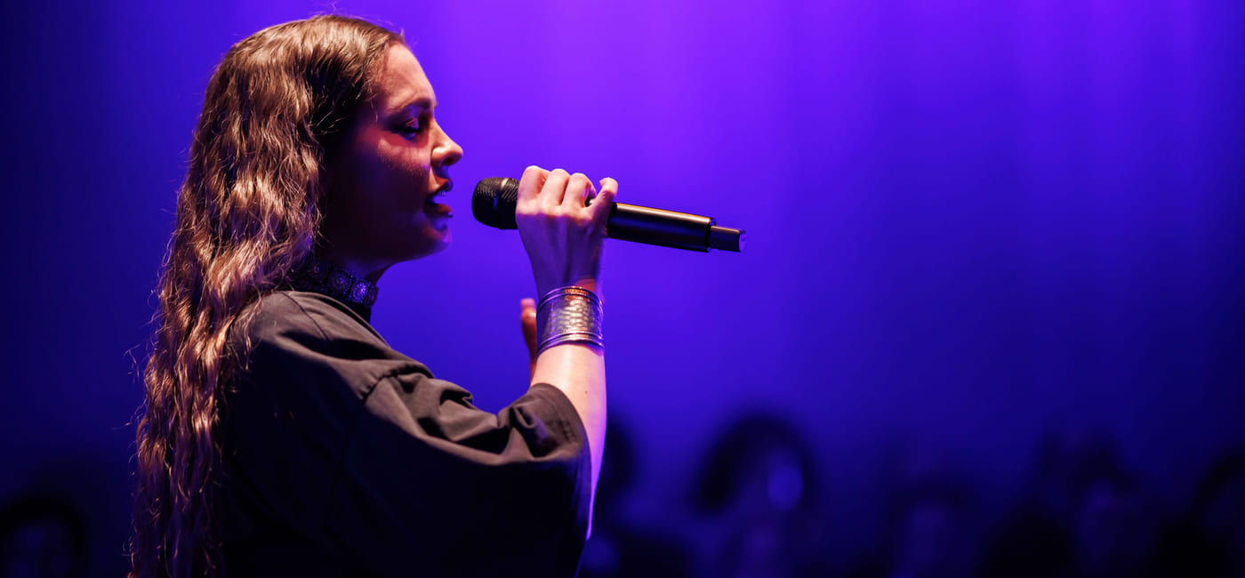 Kvinnelig sangerinne med langt bølget hår opptrer på scenen, holder mikrofonen, under lilla lys med publikumssilhuetter synlige.
