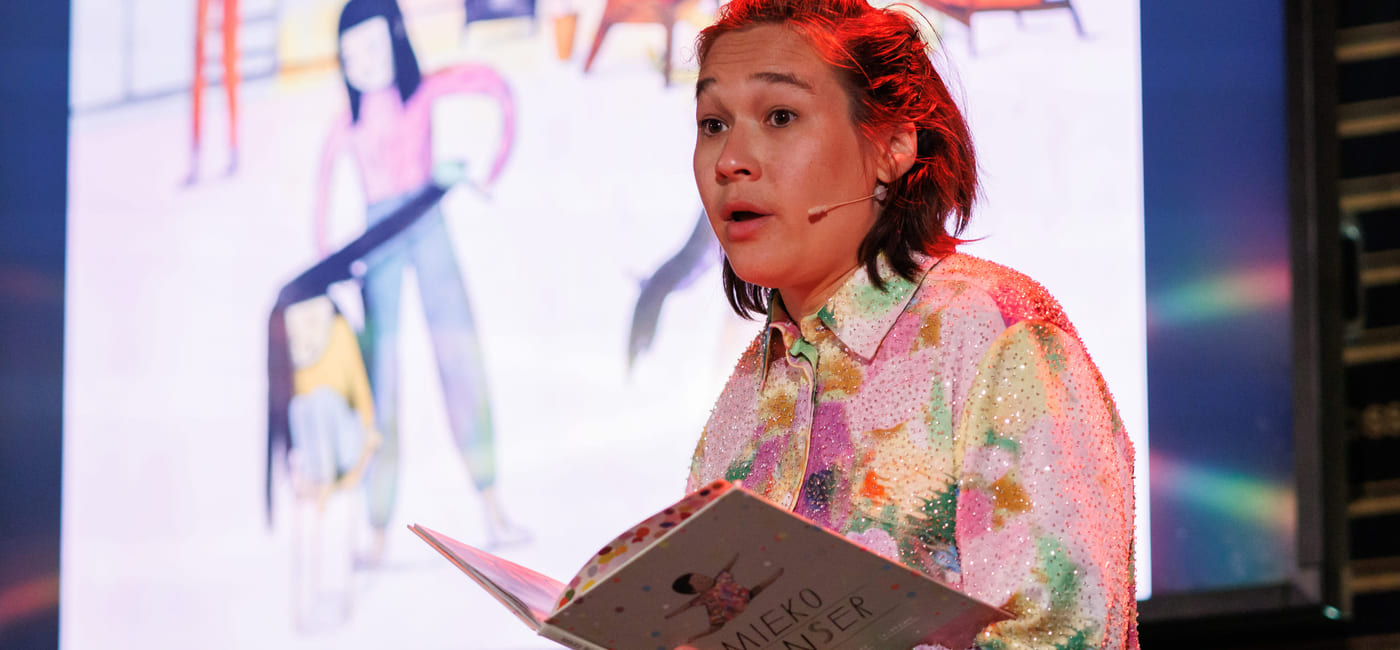 Person i fargerik paljettskjorte presenterer på scenen, holder åpen bok med tittelen Mieko Danser, med stor skjerm bak seg.