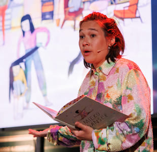 Person i fargerik paljettskjorte presenterer på scenen, holder åpen bok med tittelen Mieko Danser, med stor skjerm bak seg.