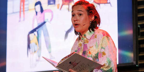 Person i fargerik paljettskjorte presenterer på scenen, holder åpen bok med tittelen Mieko Danser, med stor skjerm bak seg.
