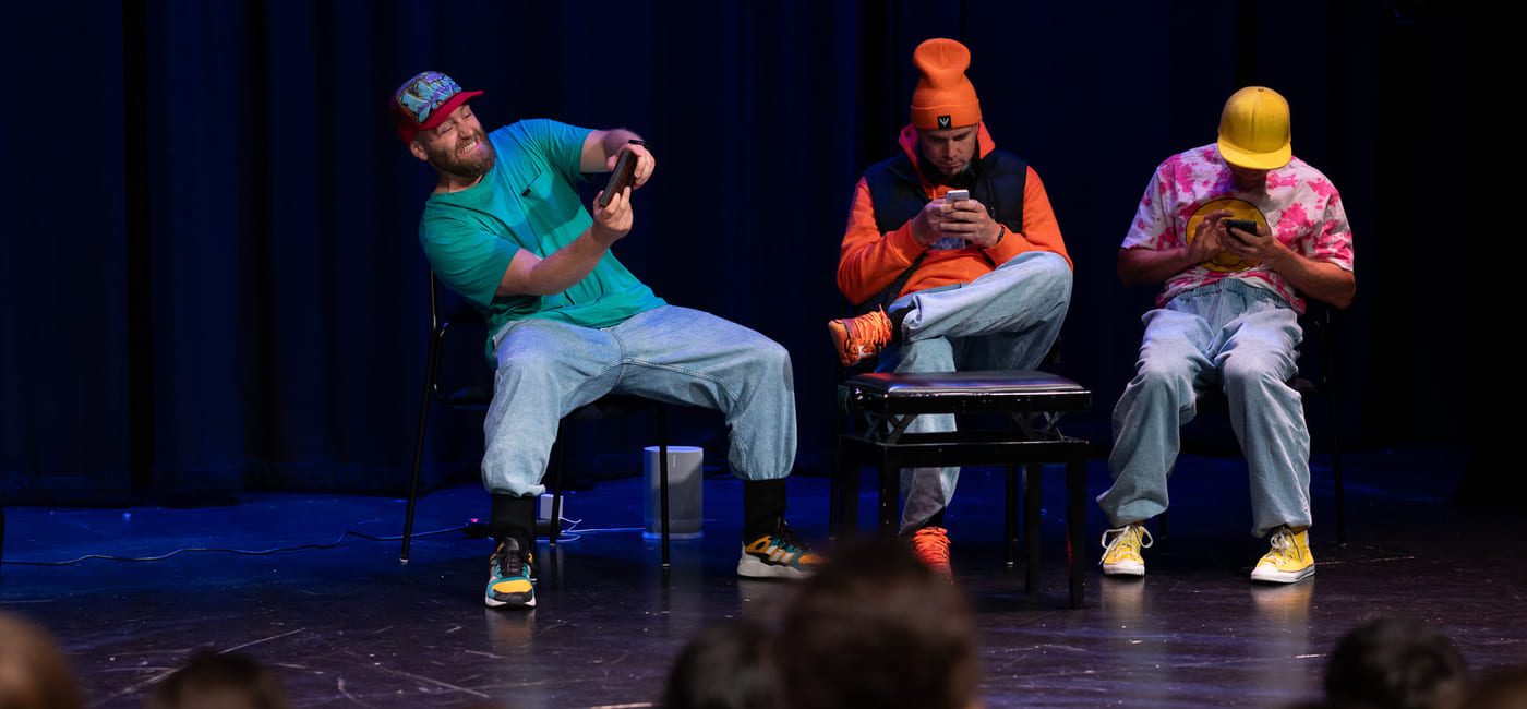 Tre menn i fargerike klær på scenen ved hjelp av smarttelefoner, med et publikum av barn synlig i forgrunnen.