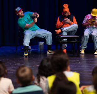 Tre menn i fargerike klær på scenen ved hjelp av smarttelefoner, med et publikum av barn synlig i forgrunnen.