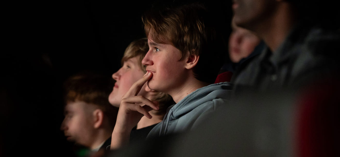 Gruppe mennesker ser intenst på i mørkt teater, ung gutt i midten med hånden på haken ser engasjert ut.