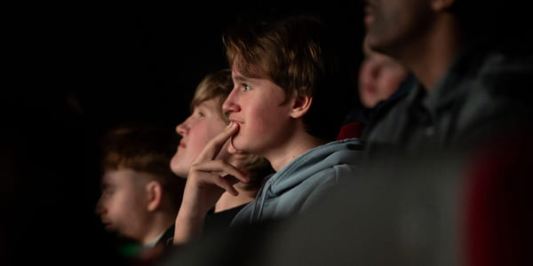 Gruppe mennesker ser intenst på i mørkt teater, ung gutt i midten med hånden på haken ser engasjert ut.