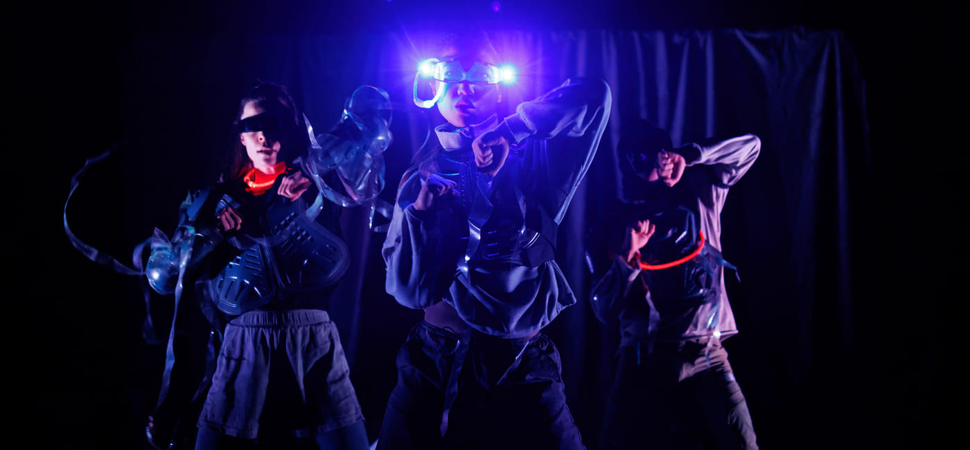 Tre artister i futuristiske kostymer danser på en mørk scene, opplyst av blått lys og glødende tilbehør.
