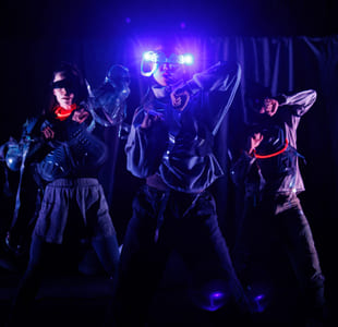 Tre artister i futuristiske kostymer danser på en mørk scene, opplyst av blått lys og glødende tilbehør.