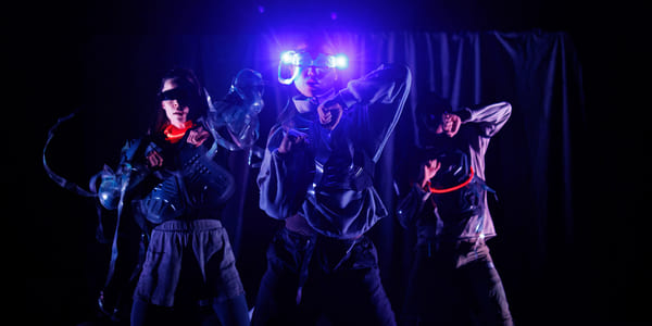 Tre artister i futuristiske kostymer danser på en mørk scene, opplyst av blått lys og glødende tilbehør.