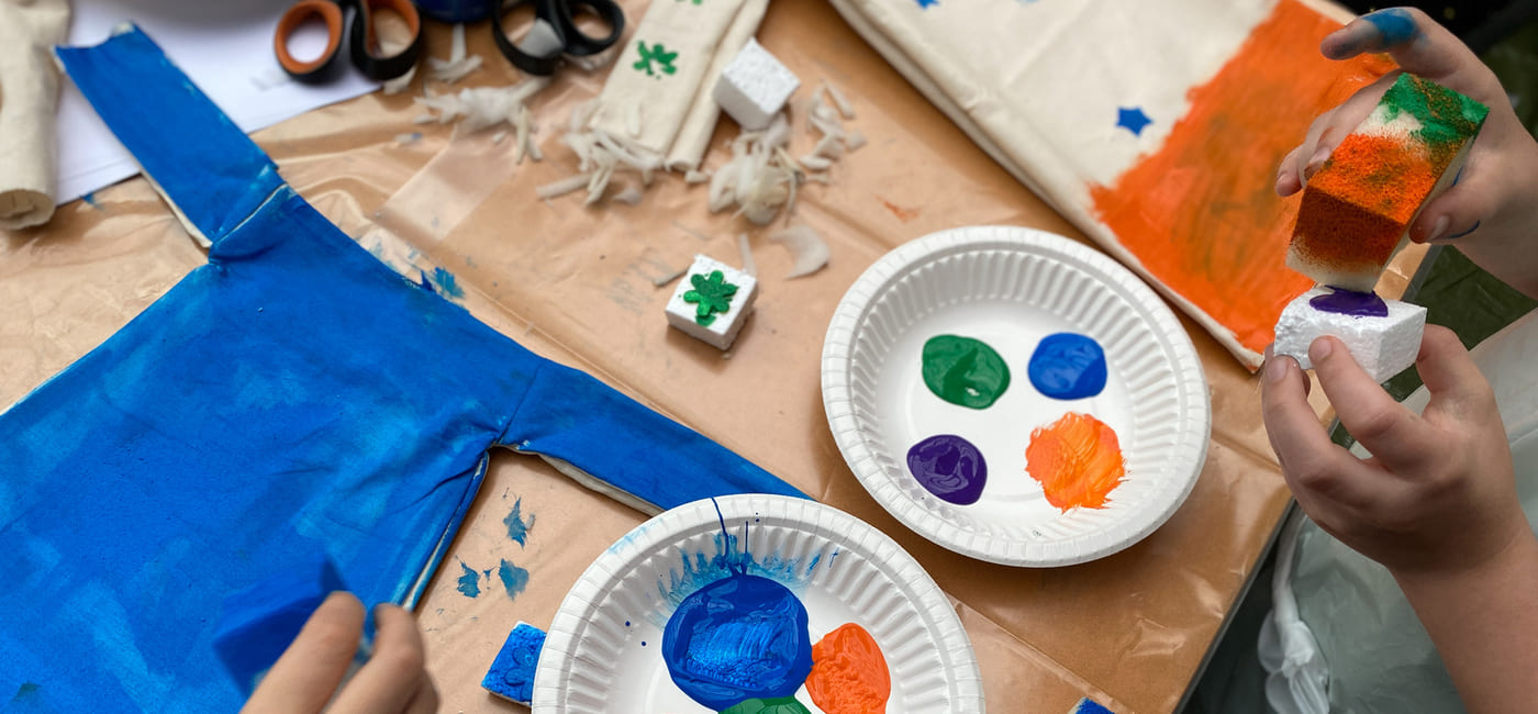 Barnehender maler med lyse farger på papirplater. Håndverksutstyr spredt på bordet, inkludert saks og blått stoff. Foto.