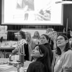 Diverse publikum ser oppmerksomt på presentasjonen i konferanserommet. Stor skjerm synlig. Bord med forfriskninger. Uformell atmosfære med engasjerte deltakere.