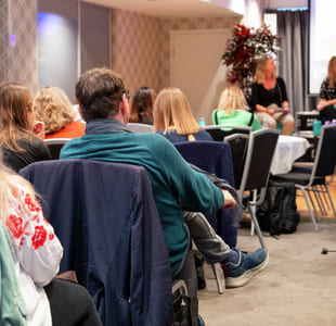 Konferansepublikum sitter vendt mot to foredragsholdere på scenen. Presentasjonsskjermer synlige. Diverse deltakere i uformelt antrekk lytter oppmerksomt.