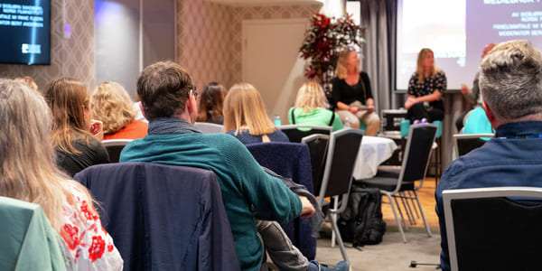 Konferansepublikum sitter vendt mot to foredragsholdere på scenen. Presentasjonsskjermer synlige. Diverse deltakere i uformelt antrekk lytter oppmerksomt.