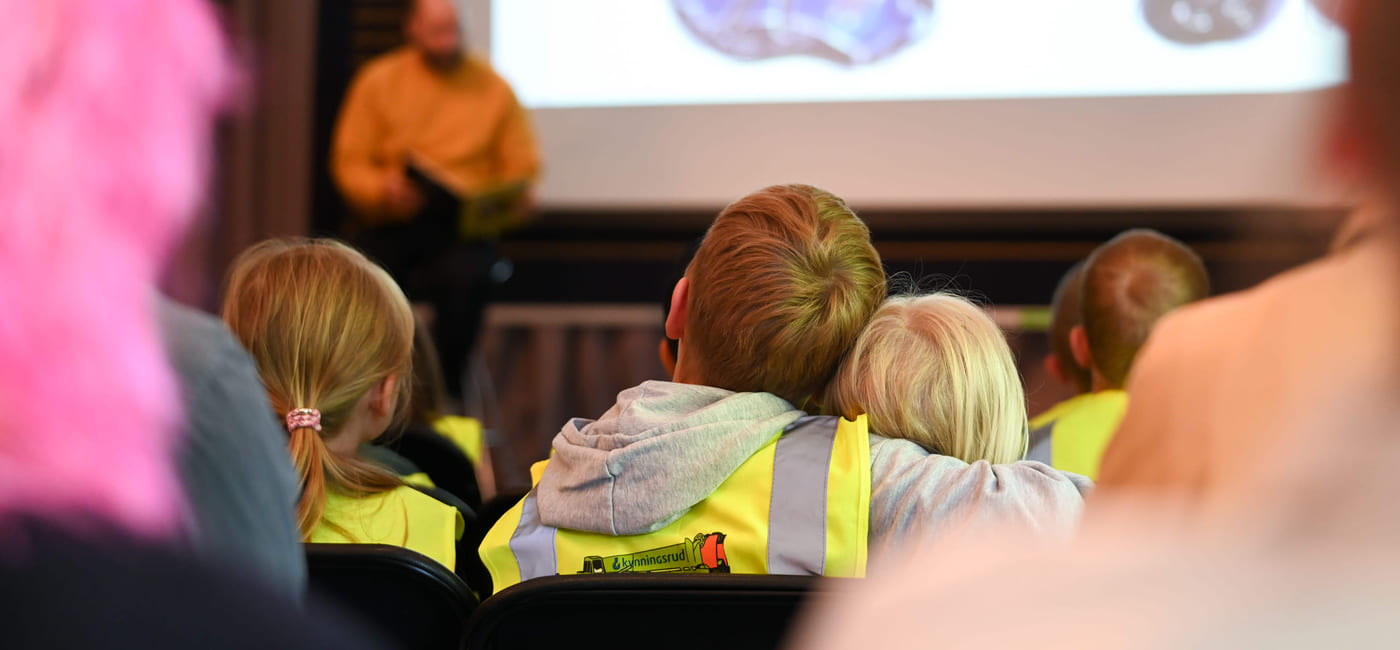 Barn i sikkerhetsvester ser på litteraturformidling. Forfatter i oransje skjorte står i nærheten av projiserte bilder av tegninger.