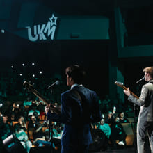To personer opptrer eller snakker foran et publikum på en scene, med scenelys og «UKM»-logo i bakgrunnen.