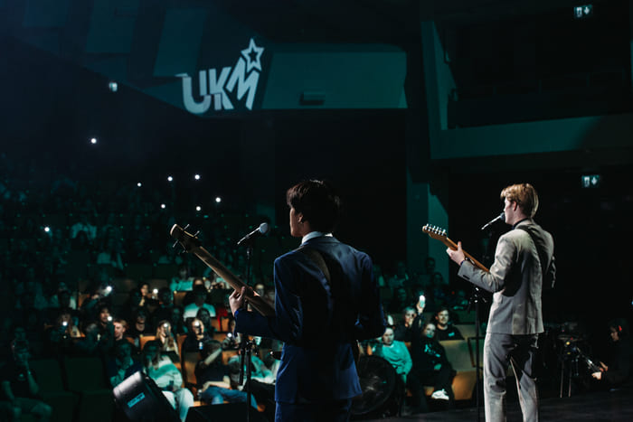 To personer opptrer eller snakker foran et publikum på en scene, med scenelys og «UKM»-logo i bakgrunnen.