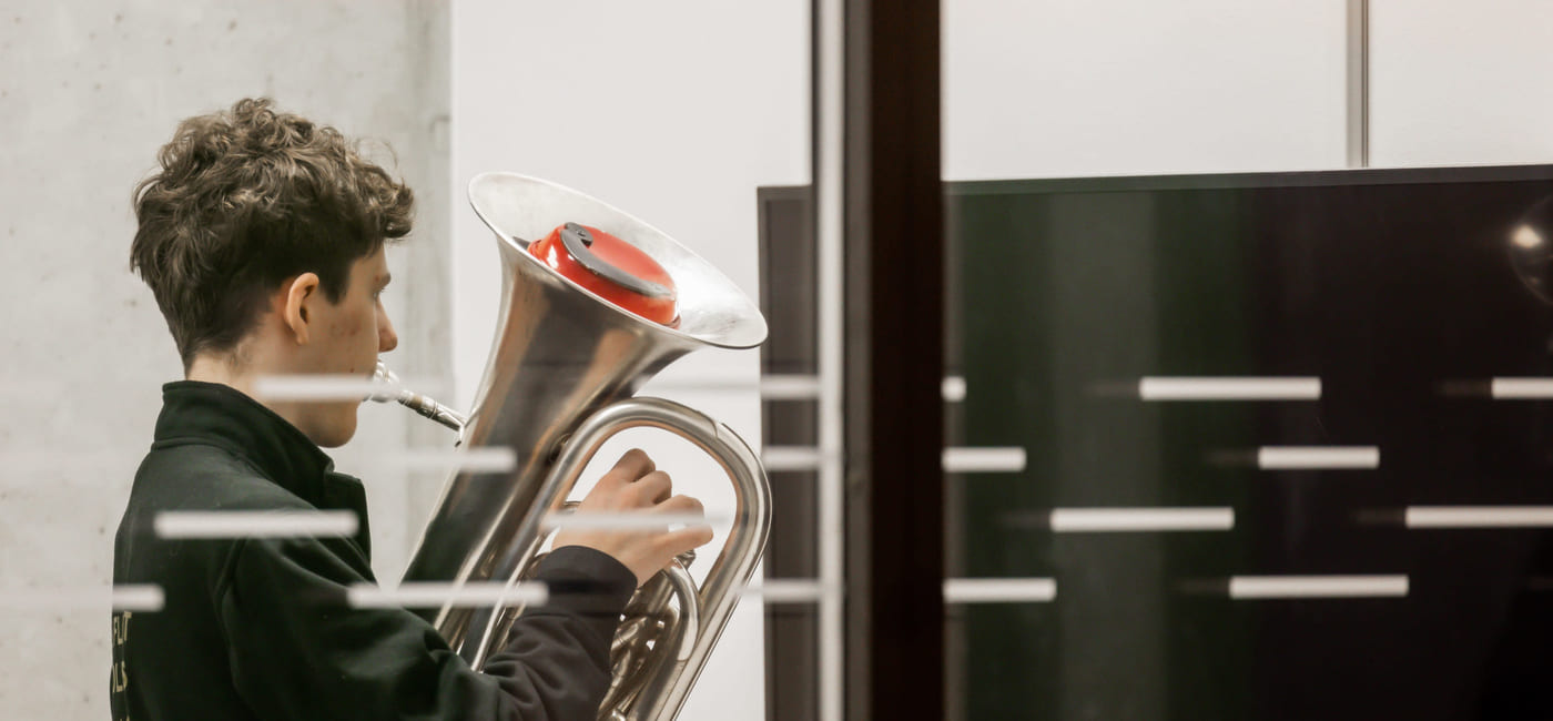 En ungdom holder en tuba og ser konsentrert ned på instrumentet i et moderne bygg.