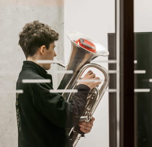 En ungdom holder en tuba og ser konsentrert ned på instrumentet i et moderne bygg.
