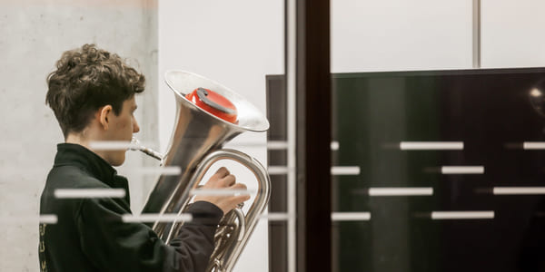 En ungdom holder en tuba og ser konsentrert ned på instrumentet i et moderne bygg.