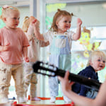 Småbarn danser i et lyst barnehagerom. En i snekkerbukse løfter gledelig armene. Fargerike dekorasjoner på vinduer. En person som spiller gitar skimtes i forgrunnen.