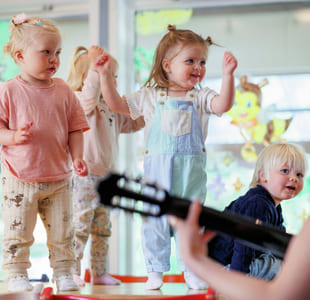 Småbarn danser i et lyst barnehagerom. En i snekkerbukse løfter gledelig armene. Fargerike dekorasjoner på vinduer. En person som spiller gitar skimtes i forgrunnen.