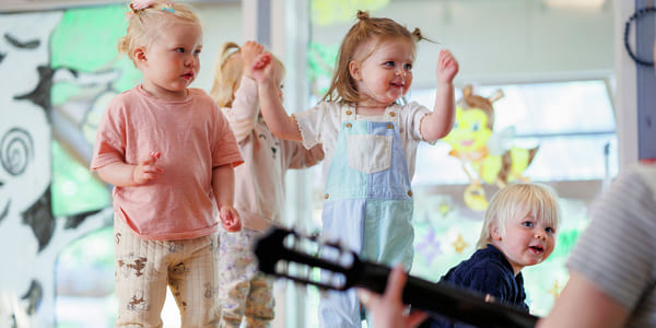 Småbarn danser i et lyst barnehagerom. En i snekkerbukse løfter gledelig armene. Fargerike dekorasjoner på vinduer. En person som spiller gitar skimtes i forgrunnen.