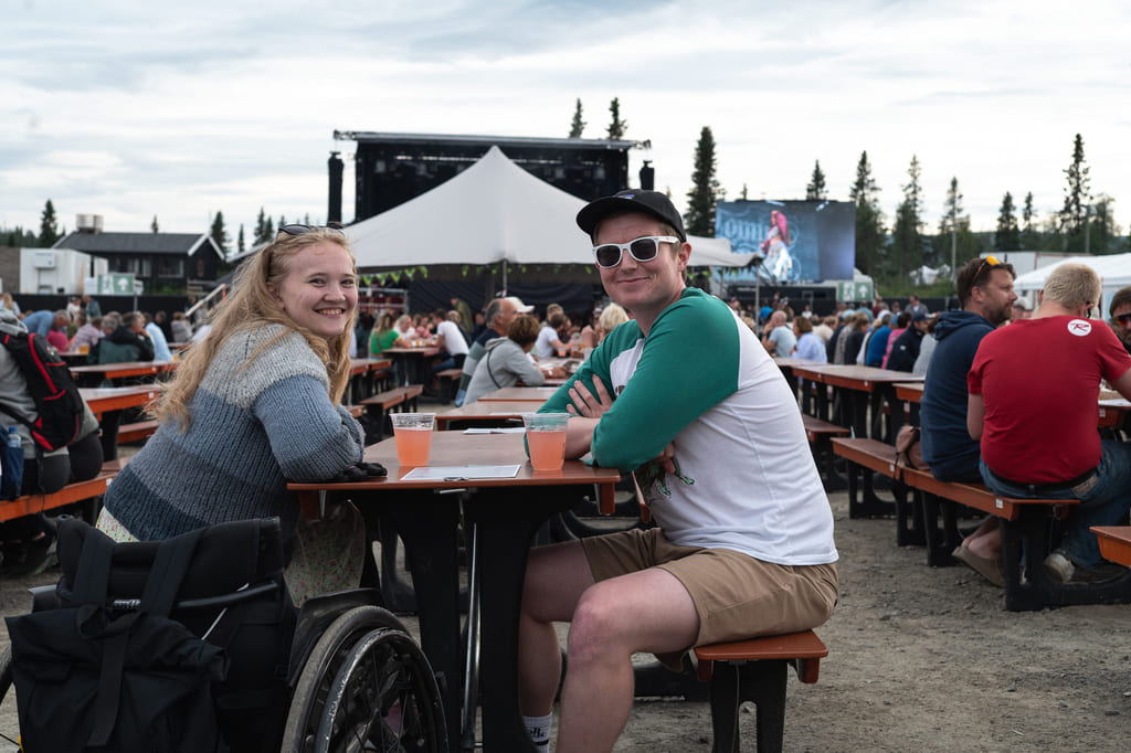 Festivalgjengere ved langbord – kvinne i rullestol og mann med caps.