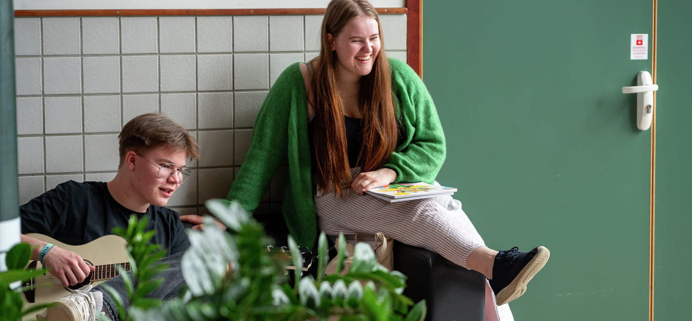 Ung mann spiller gitar, kvinne i grønn genser sitter på en avsats og smiler. Planter i forgrunnen.