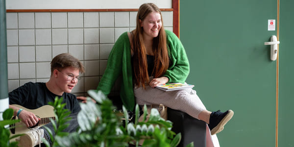 Ung mann spiller gitar, kvinne i grønn genser sitter på en avsats og smiler. Planter i forgrunnen.