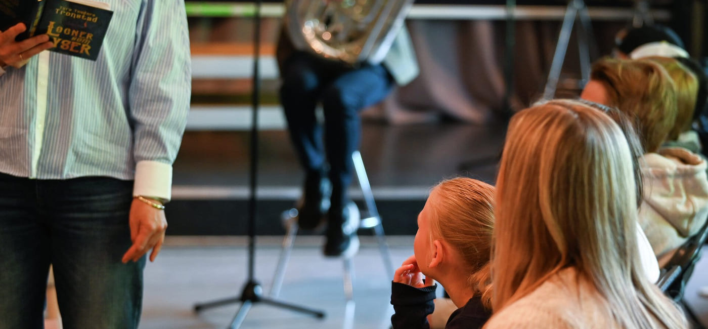 Folk som deltar på arrangementet, person som holder en bok, mikrofonstativ synlig. Publikum sitter og fokuserer på presentatøren. Uformell setting med variert belysning.