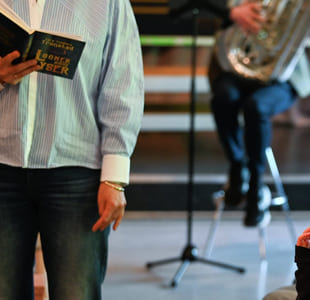 Folk som deltar på arrangementet, person som holder en bok, mikrofonstativ synlig. Publikum sitter og fokuserer på presentatøren. Uformell setting med variert belysning.