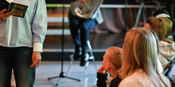 Folk som deltar på arrangementet, person som holder en bok, mikrofonstativ synlig. Publikum sitter og fokuserer på presentatøren. Uformell setting med variert belysning.