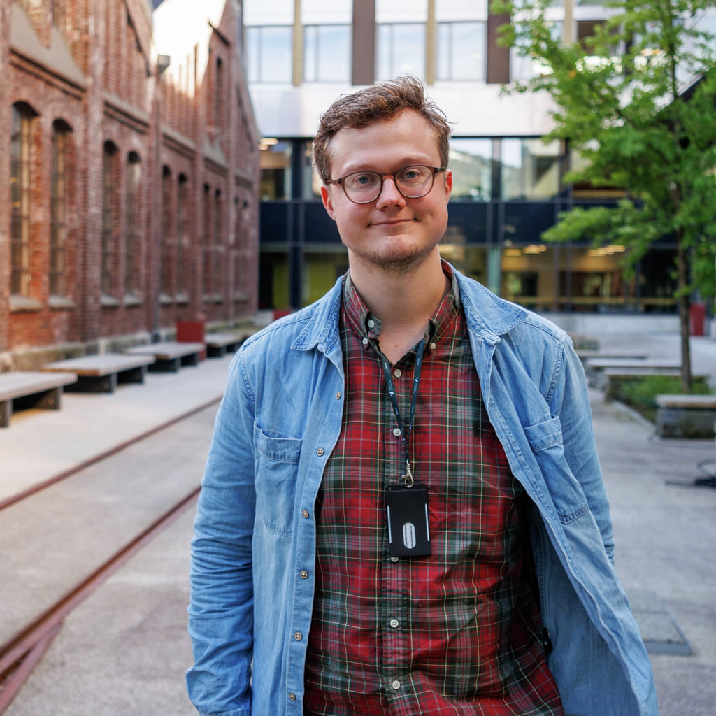 Mann i rutete skjorte og dongerijakke står i en bygård med murbygninger og benker.