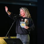 Kvinne med krøllete blondt hår som snakker i mikrofonen, holder en bok og rekker opp hånden. Iført svart genser og jeans.