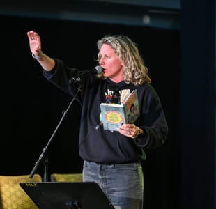 Kvinne med krøllete blondt hår som snakker i mikrofonen, holder en bok og rekker opp hånden. Iført svart genser og jeans.