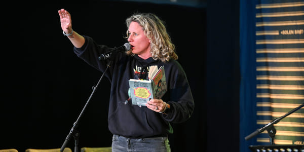 Kvinne med krøllete blondt hår som snakker i mikrofonen, holder en bok og rekker opp hånden. Iført svart genser og jeans.